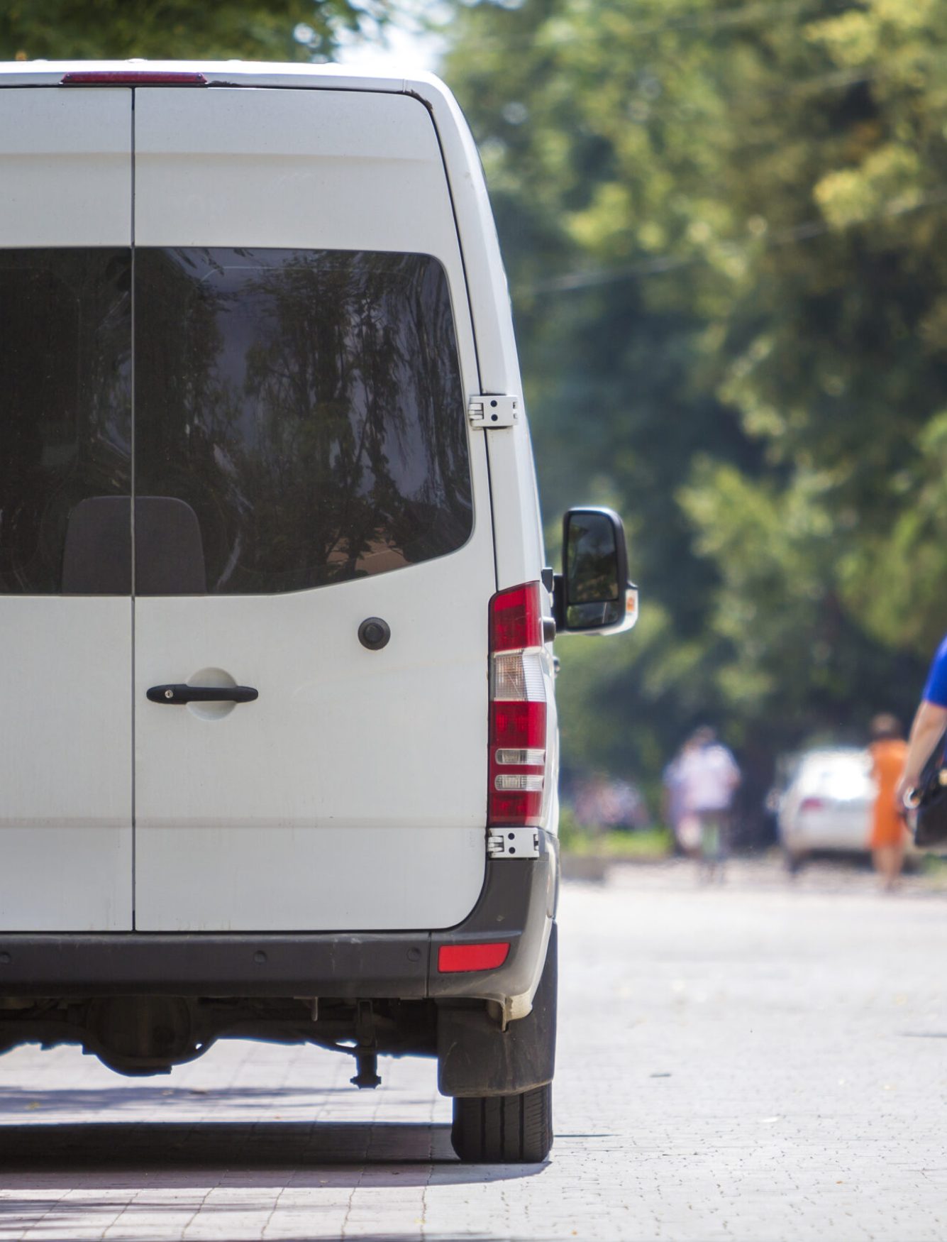 Back view of white passenger medium size commercial luxury minibus van parked n shadow of green tree on summer city street i with blurred silhouettes of pedestrians and cars under green trees.